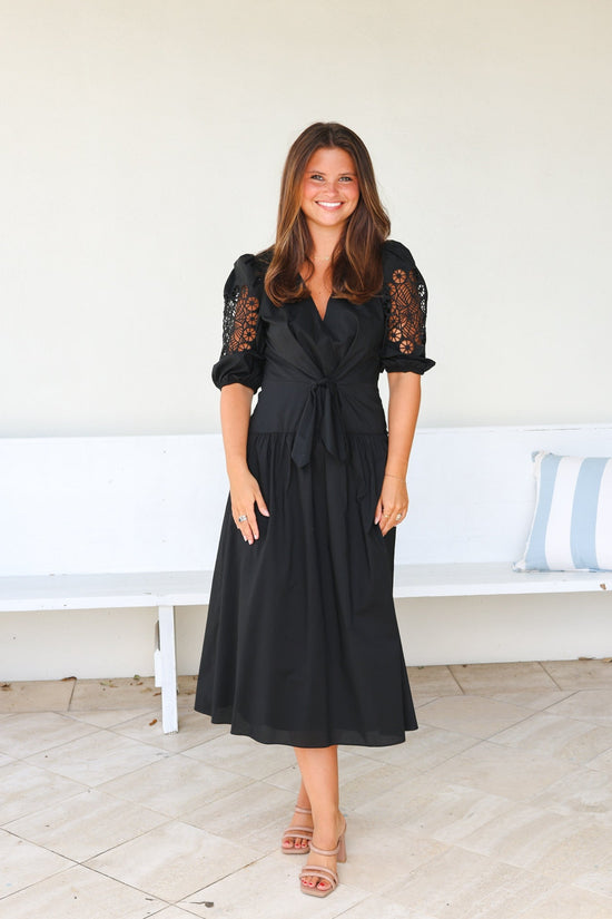 Woman wearing a black dress with lace sleeves standing in a room with a white bench and striped pillows.